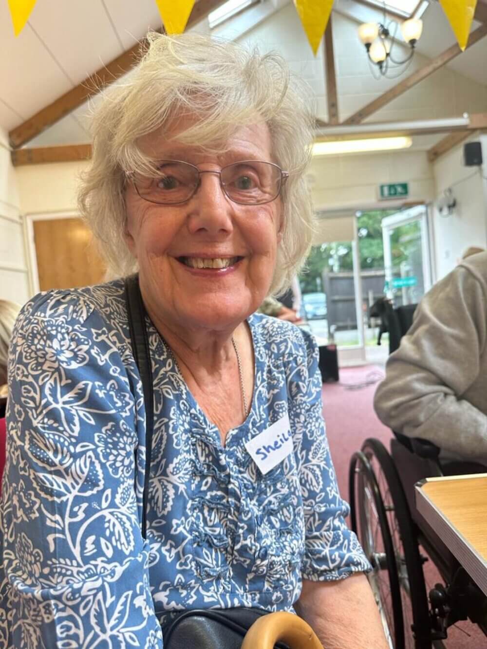 An elderly woman with glasses and a blue floral shirt smiling. She has a name tag reading "Sheila. - Home Instead