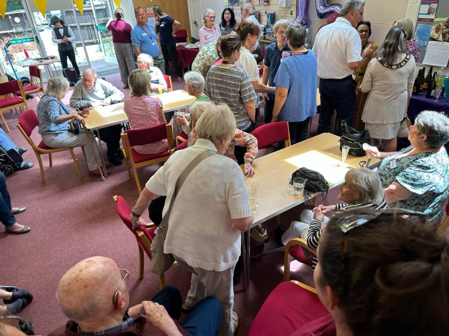 A group of elderly people socializing in a community center, with some seated at tables and others standing and mingling. - Home Instead