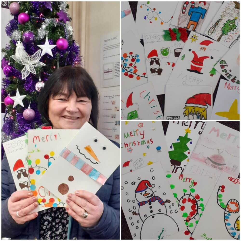 A woman smiling and holding holiday cards in front of a decorated Christmas tree. Many holiday cards are displayed nearby. - Home Instead