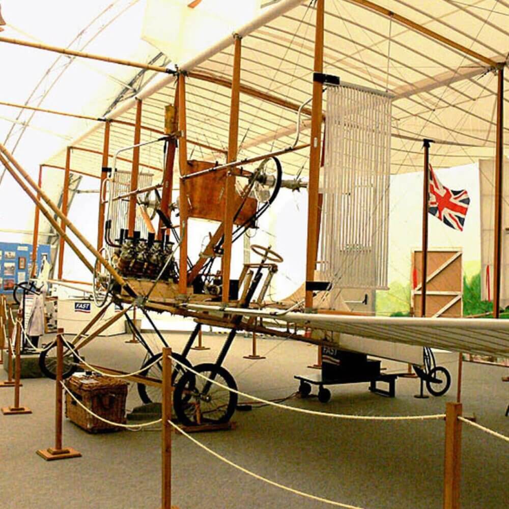 Vintage aircraft with open cockpit and exposed mechanics displayed in a museum, with a British flag in the background. - Home Instead