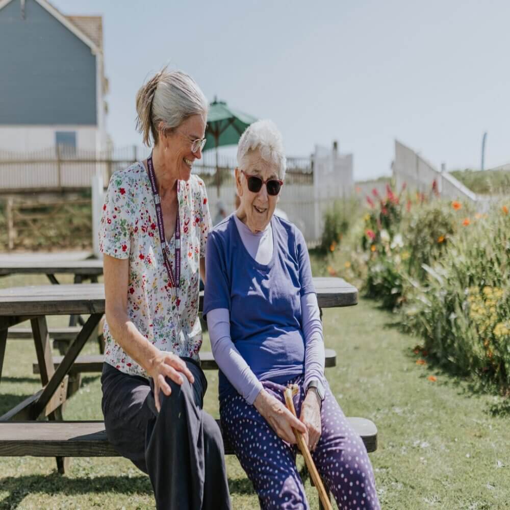 Two women sitting on a bench outdoors, smiling and engaged in conversation on a sunny day. - Home Instead