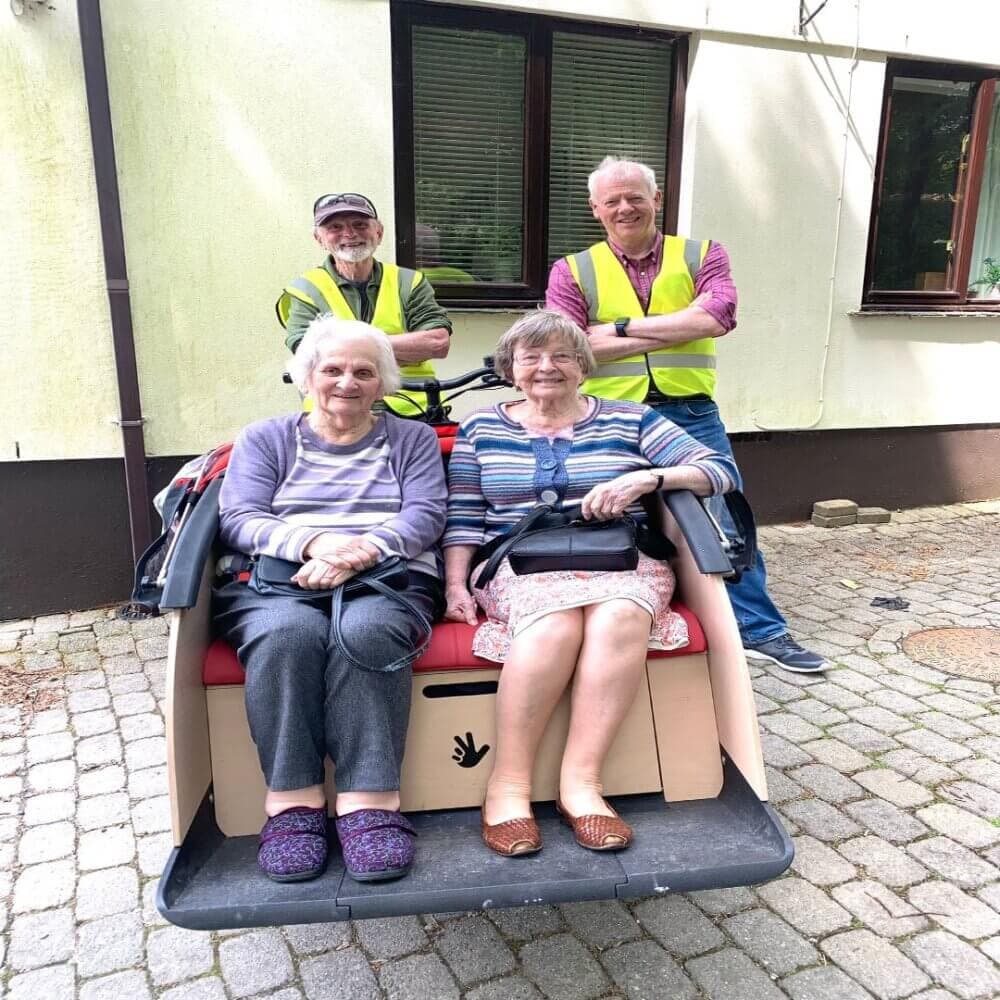 Two elderly women sit side by side in a trishaw while two men in high-visibility vests stand behind them, smiling. - Home Instead