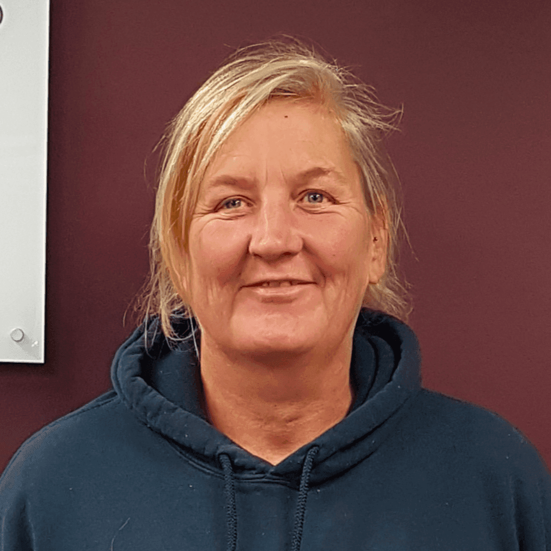 A person with blonde hair in a blue hoodie smiles against a maroon background. - Home Instead