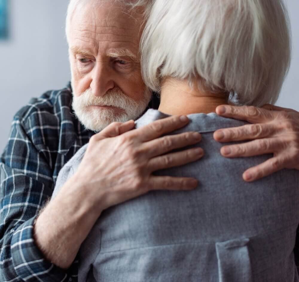 An elderly man with white hair and a beard hugs an elderly woman with short white hair, both appearing emotional. - Home Instead