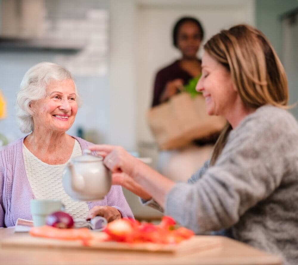 An elderly woman and a younger woman smiling, sitting at a table with food, while another person stands in the background. - Home Instead