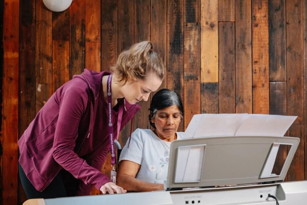 A young woman assists an older woman playing an electronic keyboard against a wooden wall background. - Home Instead
