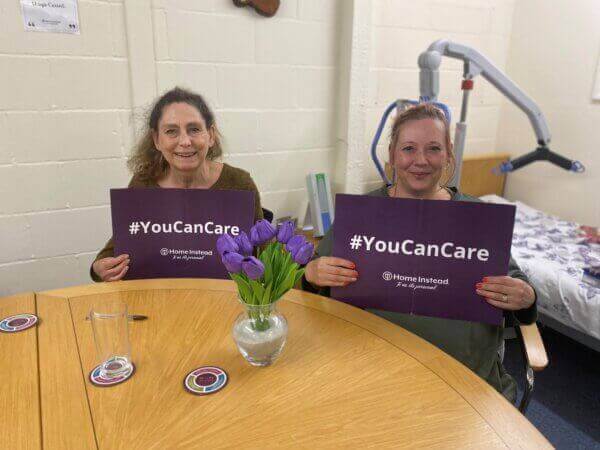 Two people at a table hold "#YouCanCare" signs. A vase of purple flowers is on the table between them. - Home Instead