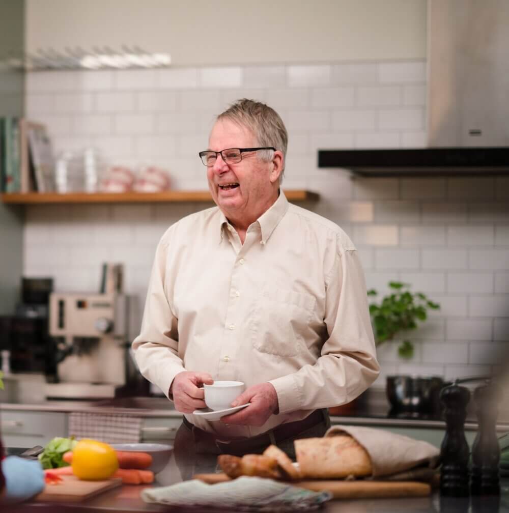 Elderly man holding a cup and smiling in a kitchen, with various food items on the counter in front of him. - Home Instead