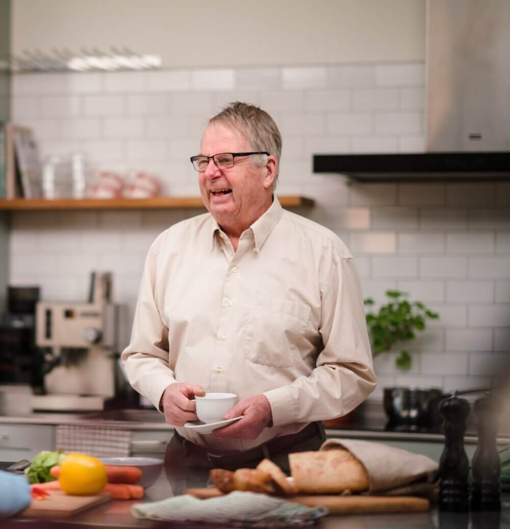 Smiling elderly man holding teacup in modern kitchen, with bread, vegetables, and kitchen appliances in the background. - Home Instead
