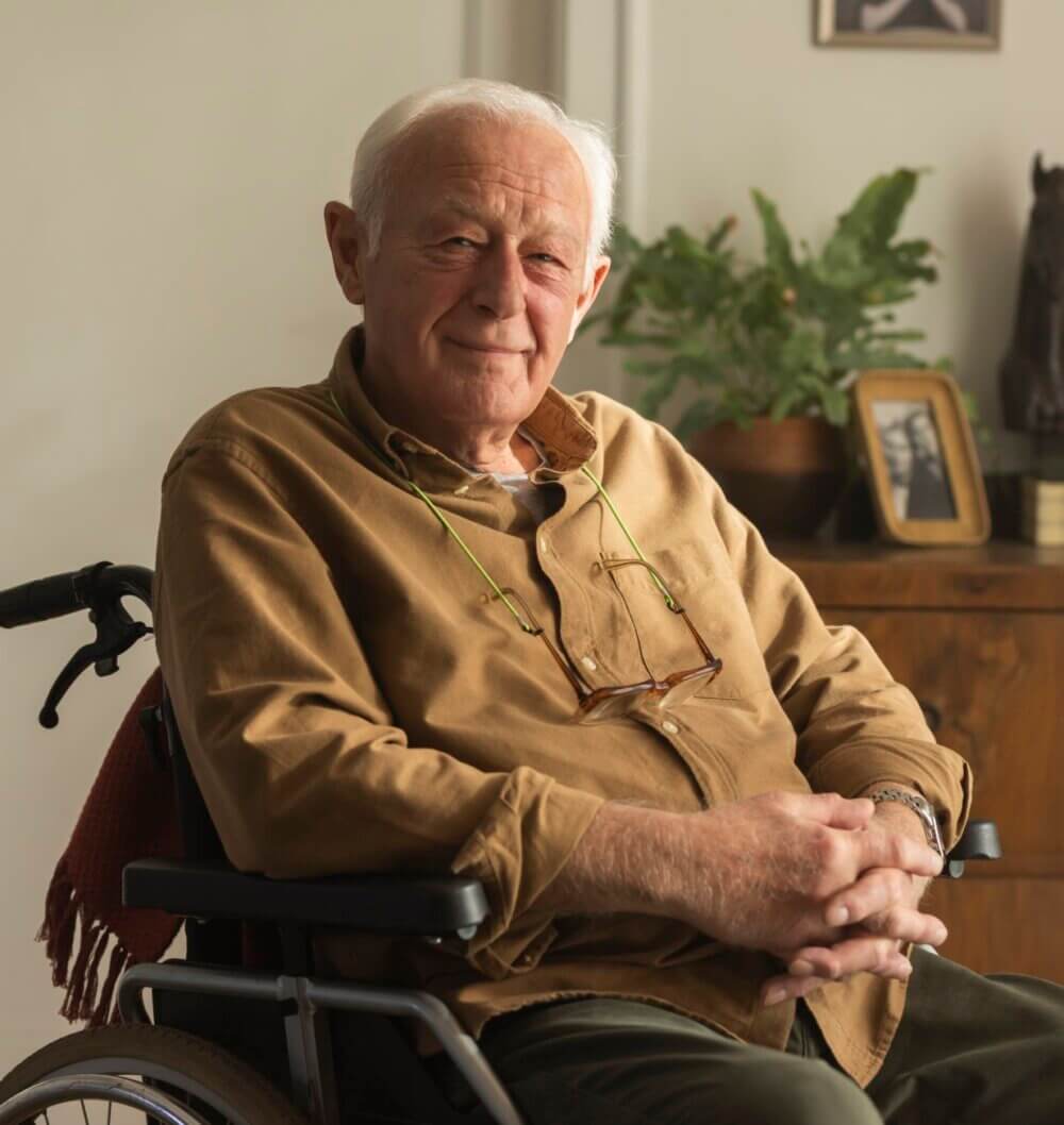 An elderly man with a warm smile sits in a wheelchair, wearing a brown shirt. A plant and framed photos are in the background. - Home Instead