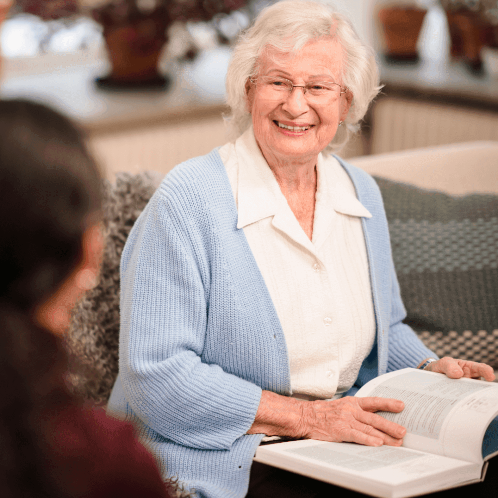 Elderly woman with glasses wearing a light blue cardigan, smiling, and holding an open book, sitting indoors. - Home Instead