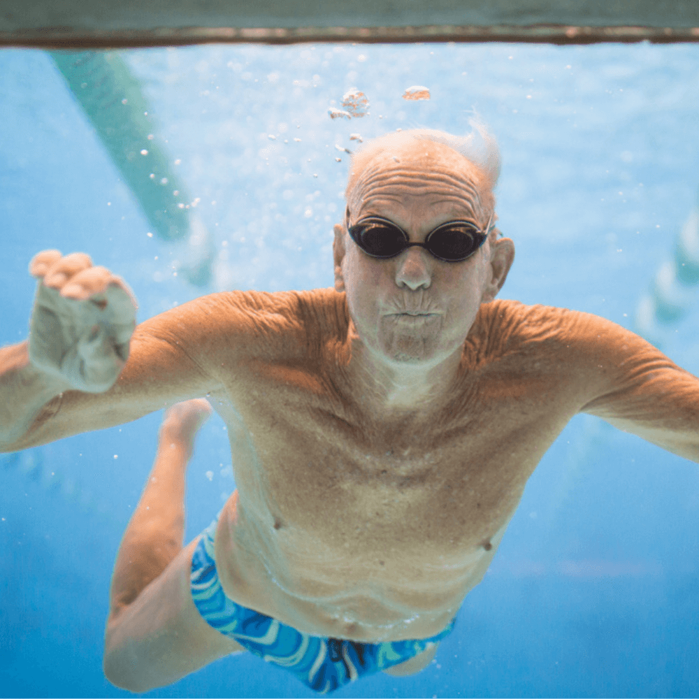 Elderly man swimming underwater in a pool, wearing blue swim trunks and black goggles. - Home Instead