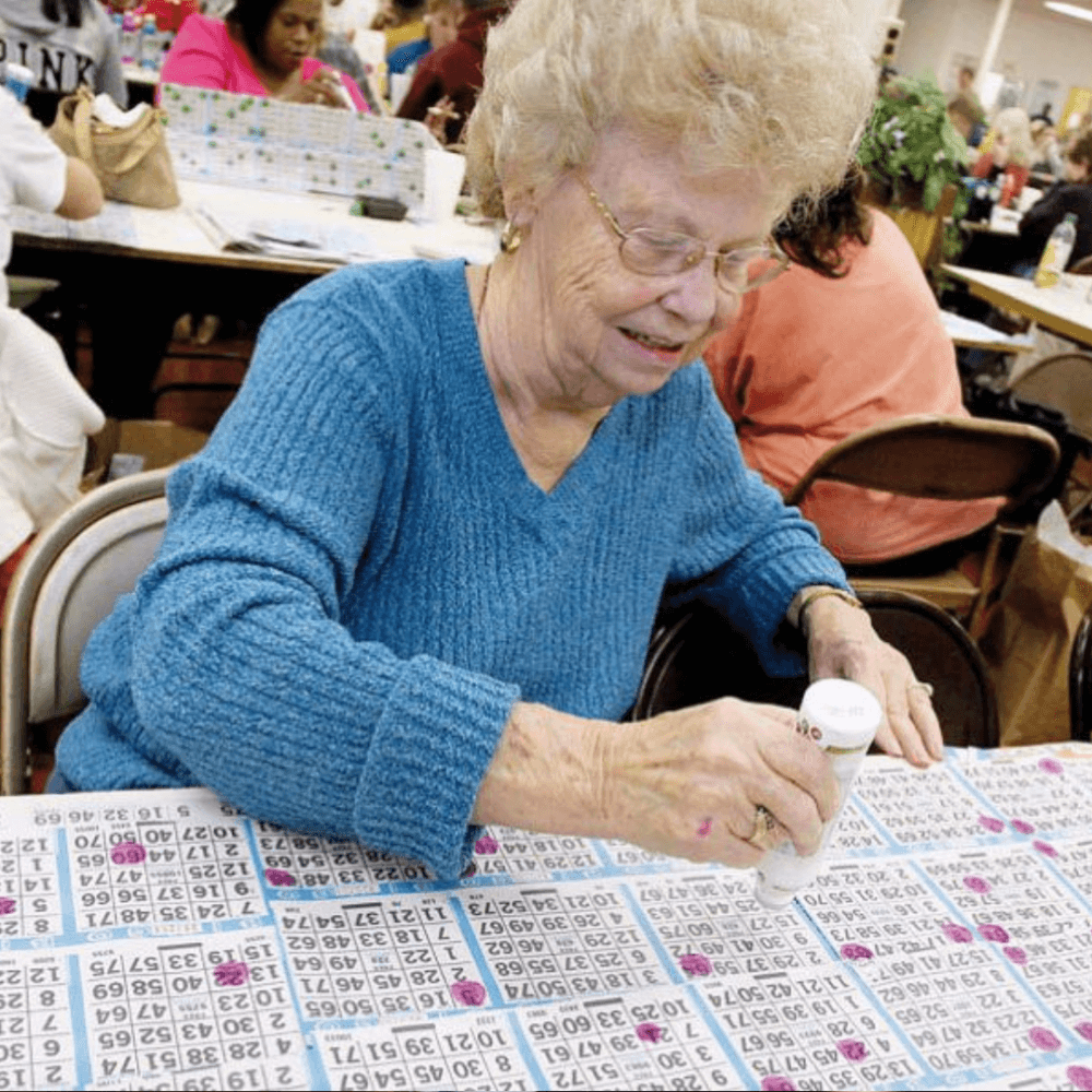 A woman with short curly hair and glasses is playing bingo, using a dabber on her bingo cards. - Home Instead