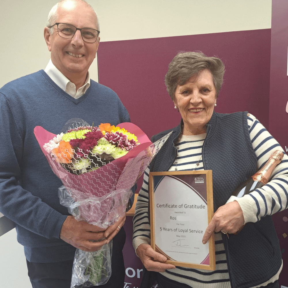 Two people smiling; one holds a bouquet, and the other displays a certificate of gratitude for 5 years of loyal service. - Home Instead