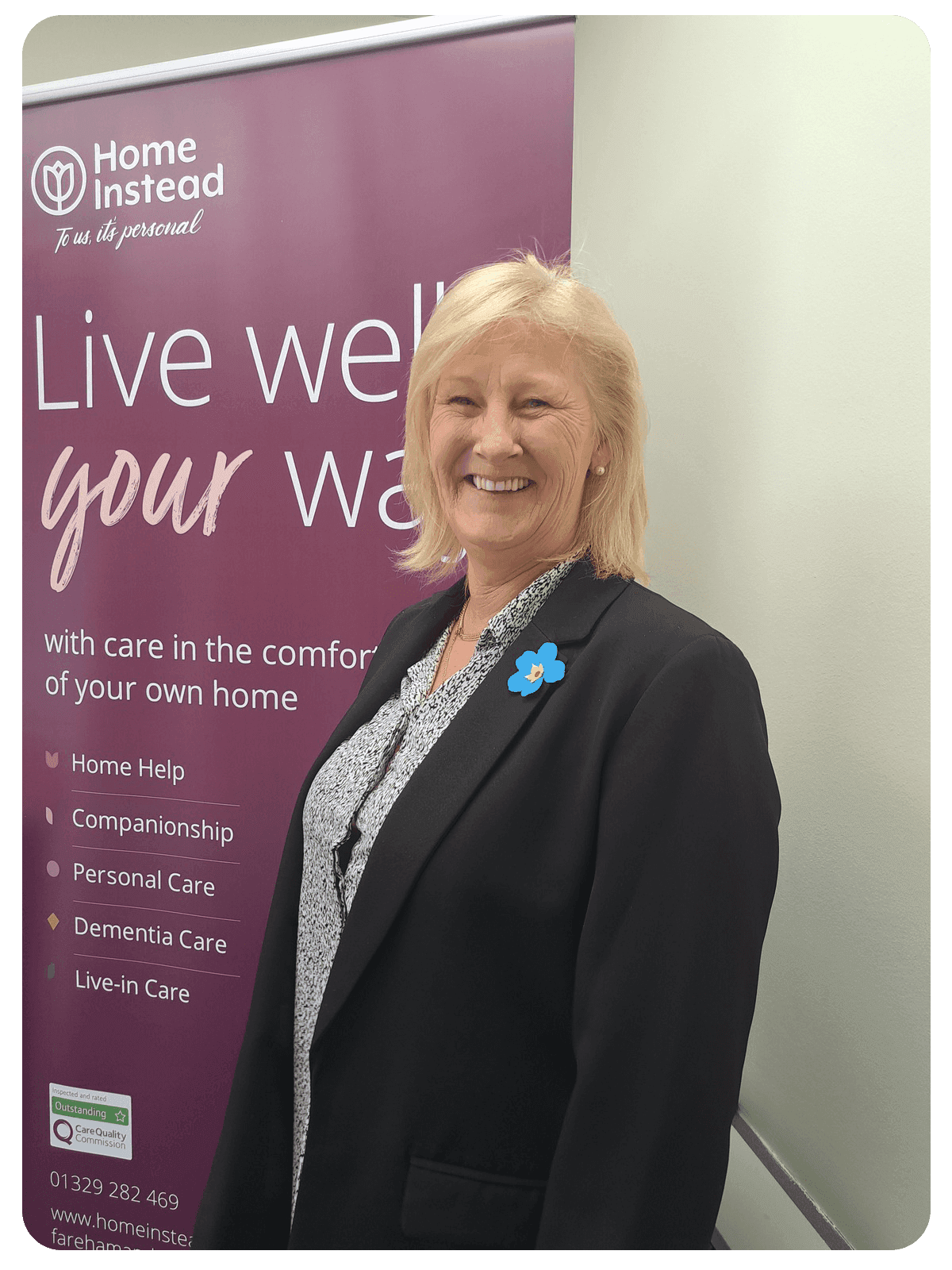 A woman in a black blazer stands smiling in front of a Home Instead banner with care service details. - Home Instead