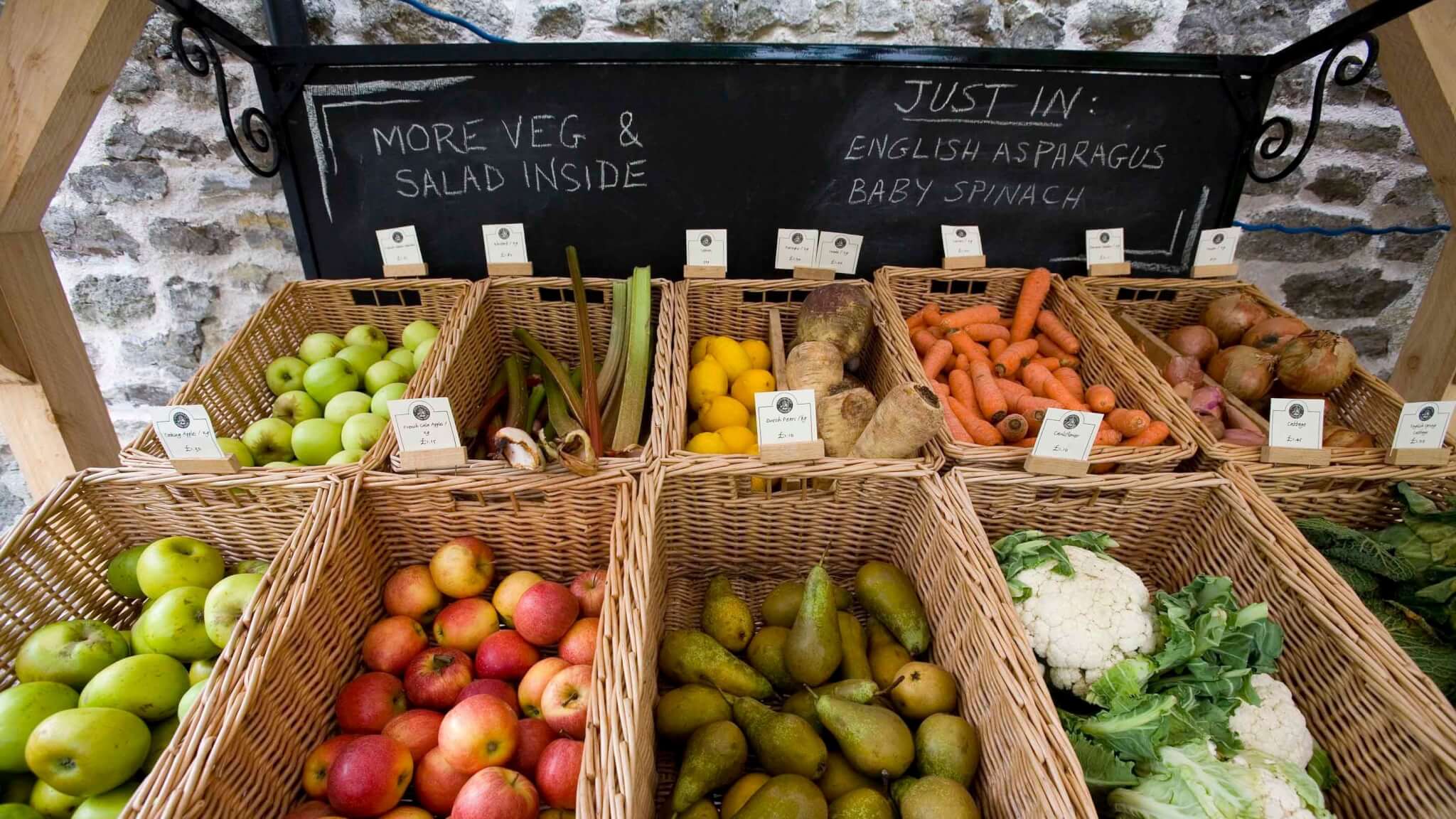 Basket display of fresh fruits and vegetables at a market, with labels and a chalkboard sign listing new arrivals. - Home Instead