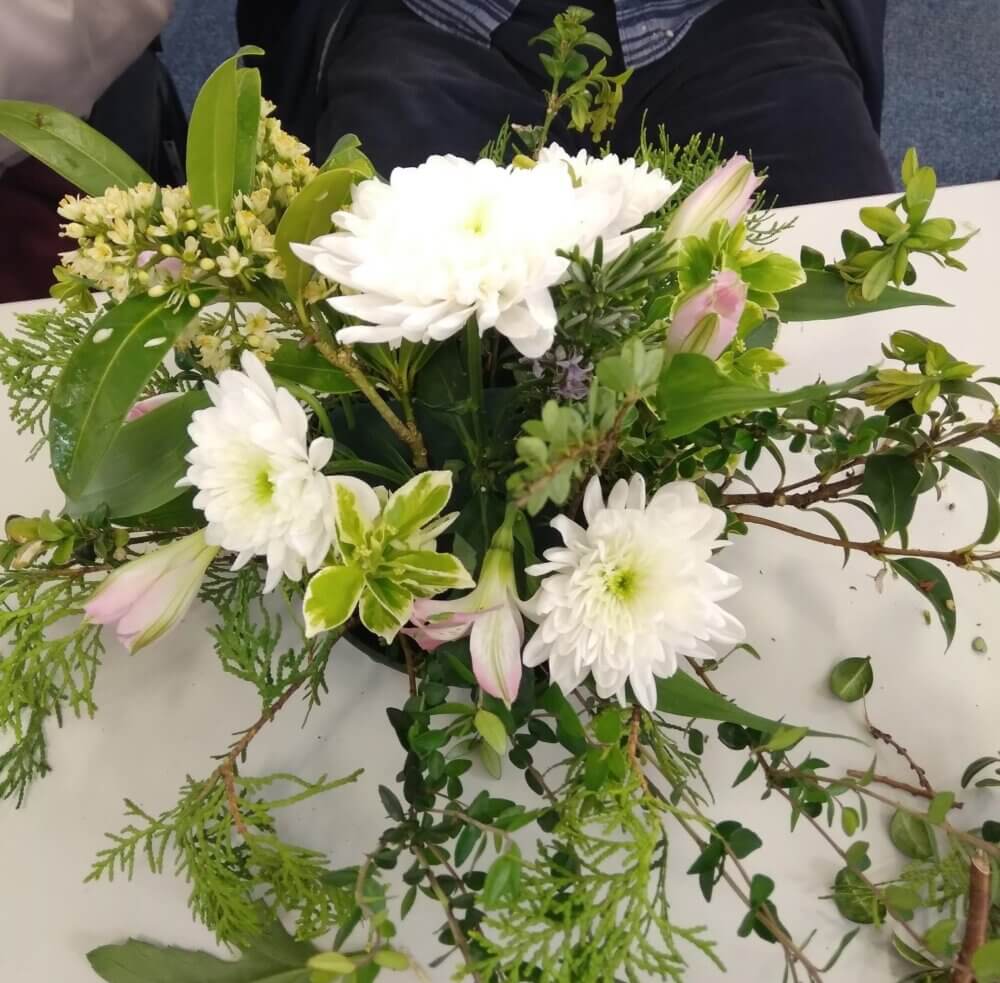 A floral arrangement featuring white flowers and green foliage sits on a table. - Home Instead