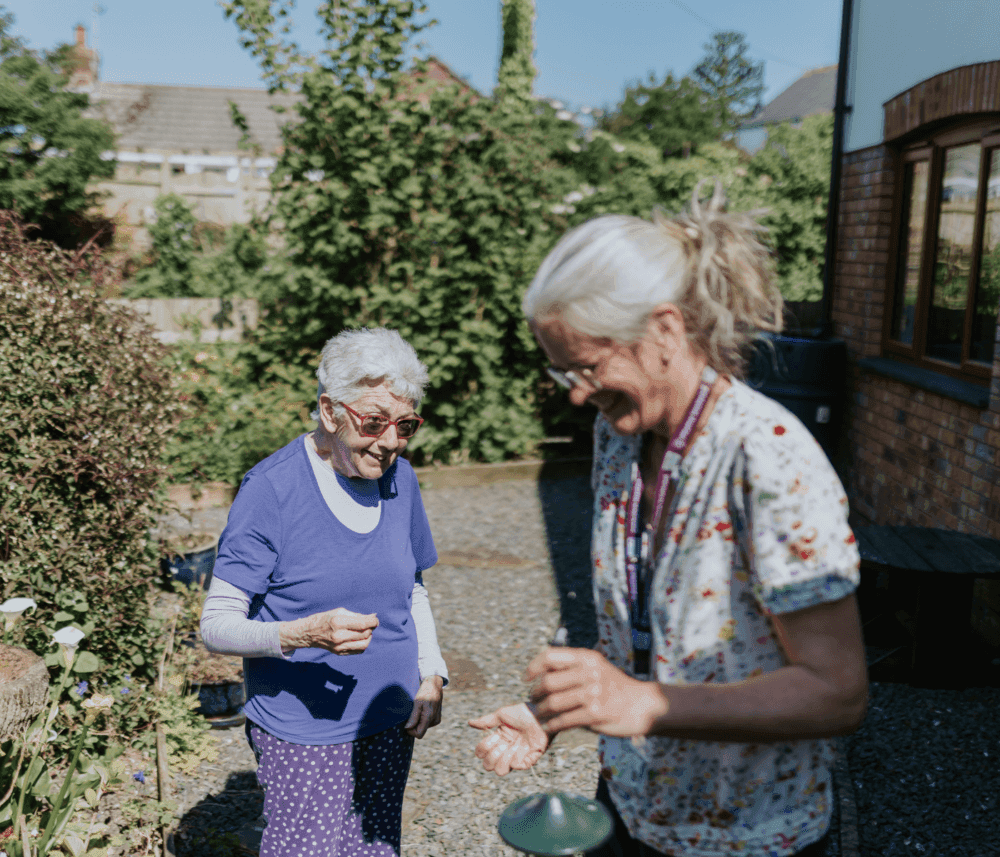 Two elderly women enjoy a sunny day outside, laughing together in a garden. - Home Instead