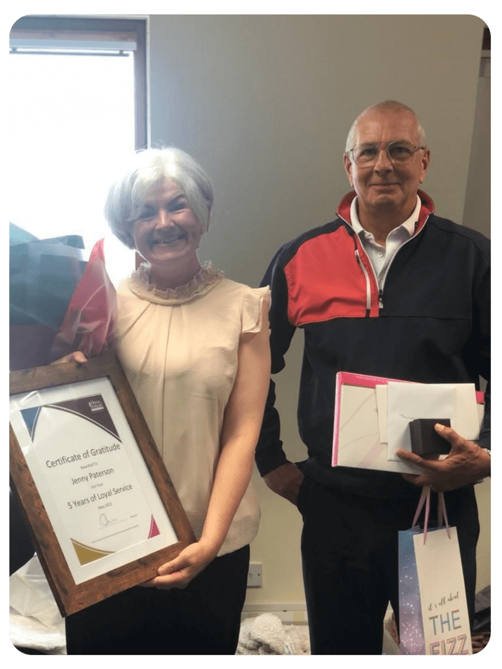 Two people posing, the woman holding a "Certificate of Gratitude" and a bouquet, both smiling. - Home Instead