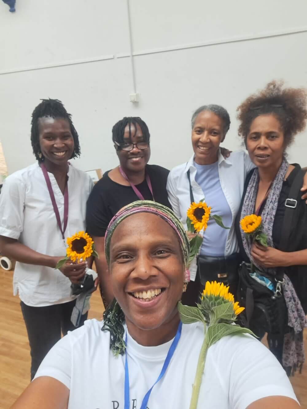 Five smiling people holding sunflowers and standing together in a room with wooden floors and a white wall. - Home Instead