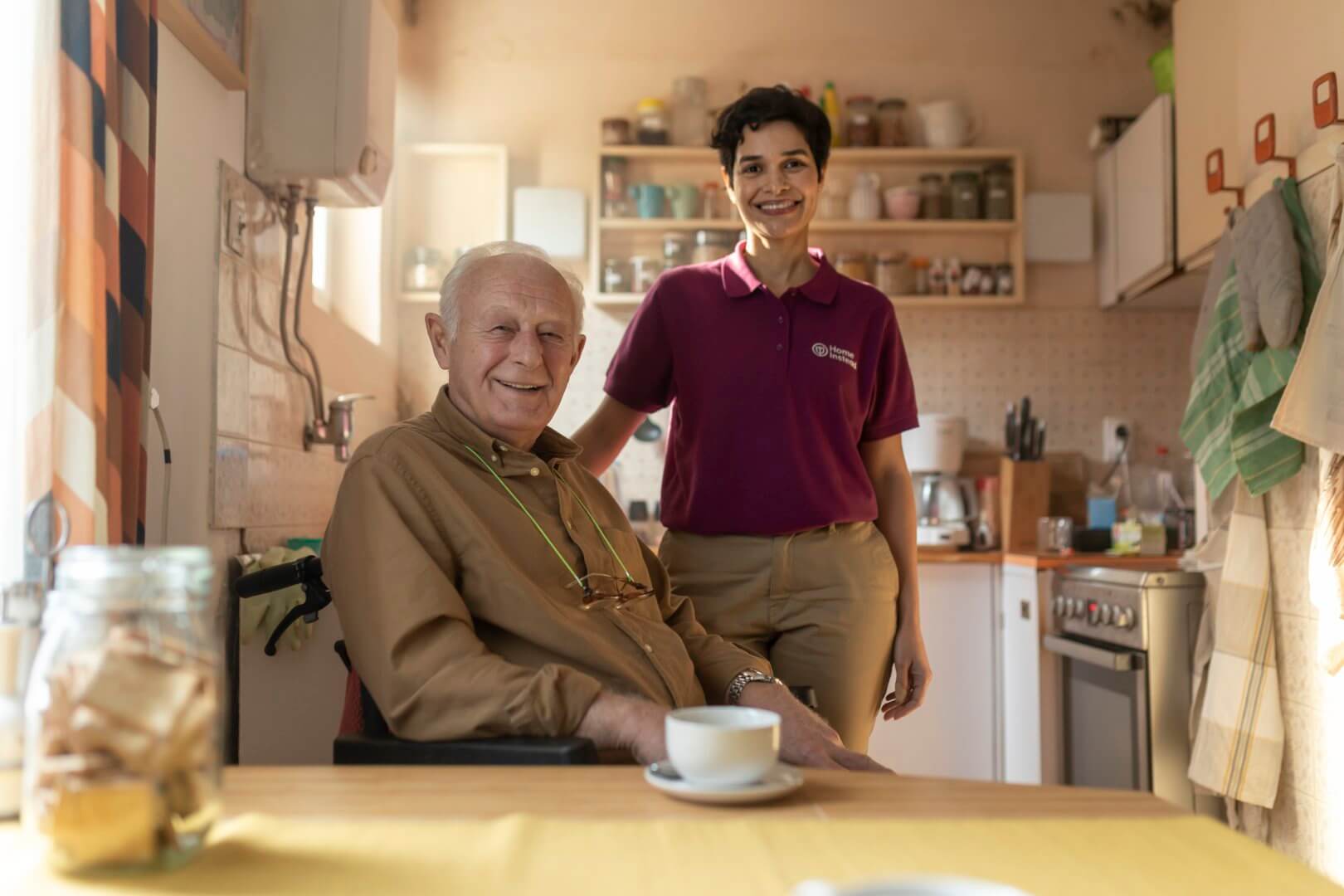 An elderly man in a wheelchair smiles while a caregiver stands beside him in a cozy kitchen. - Home Instead