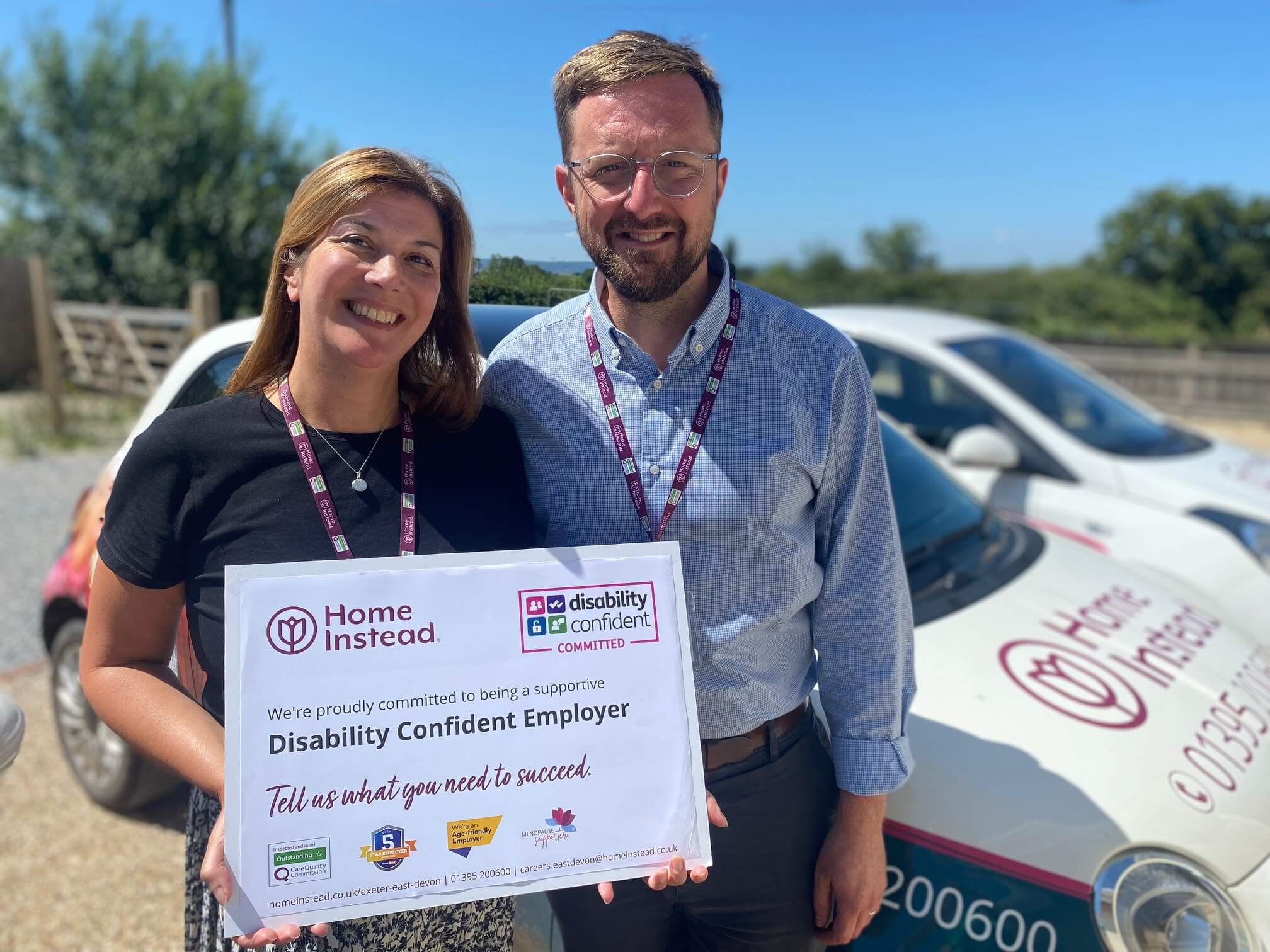 Two people hold a "Disability Confident Employer" sign, standing in front of a car with "Home Instead" branding. - Home Instead