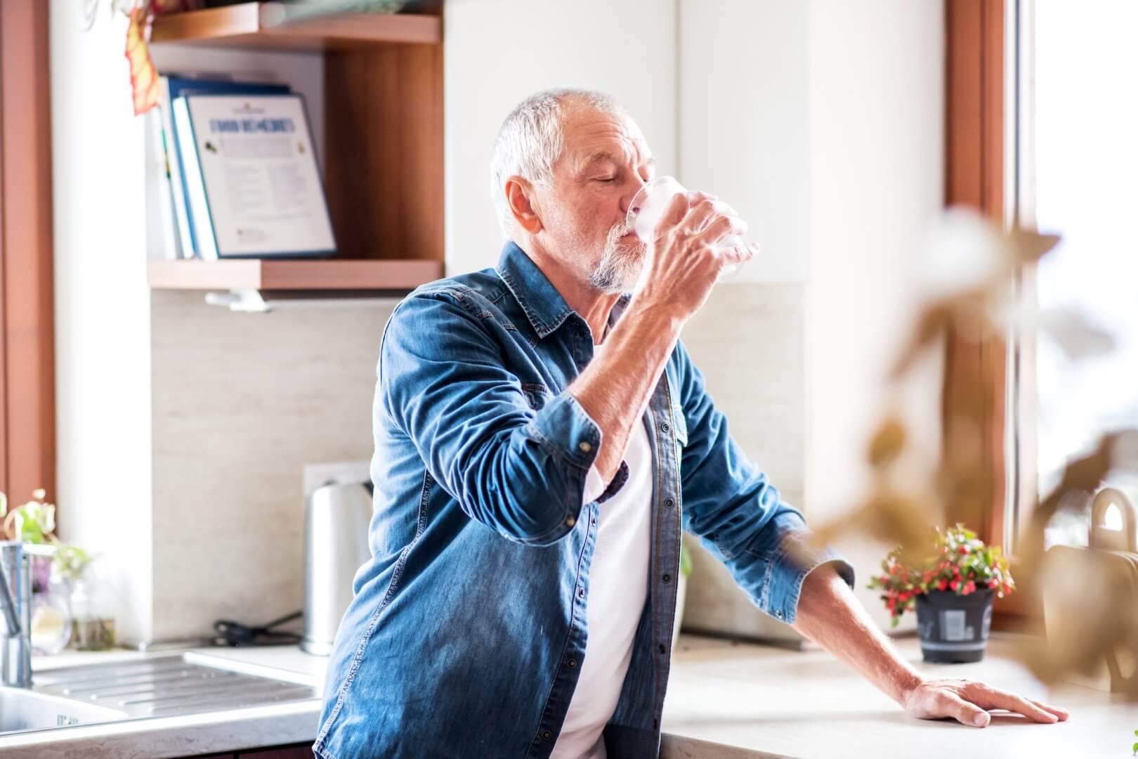 Elderly man in a denim shirt drinks water in a bright kitchen near a window. - Home Instead