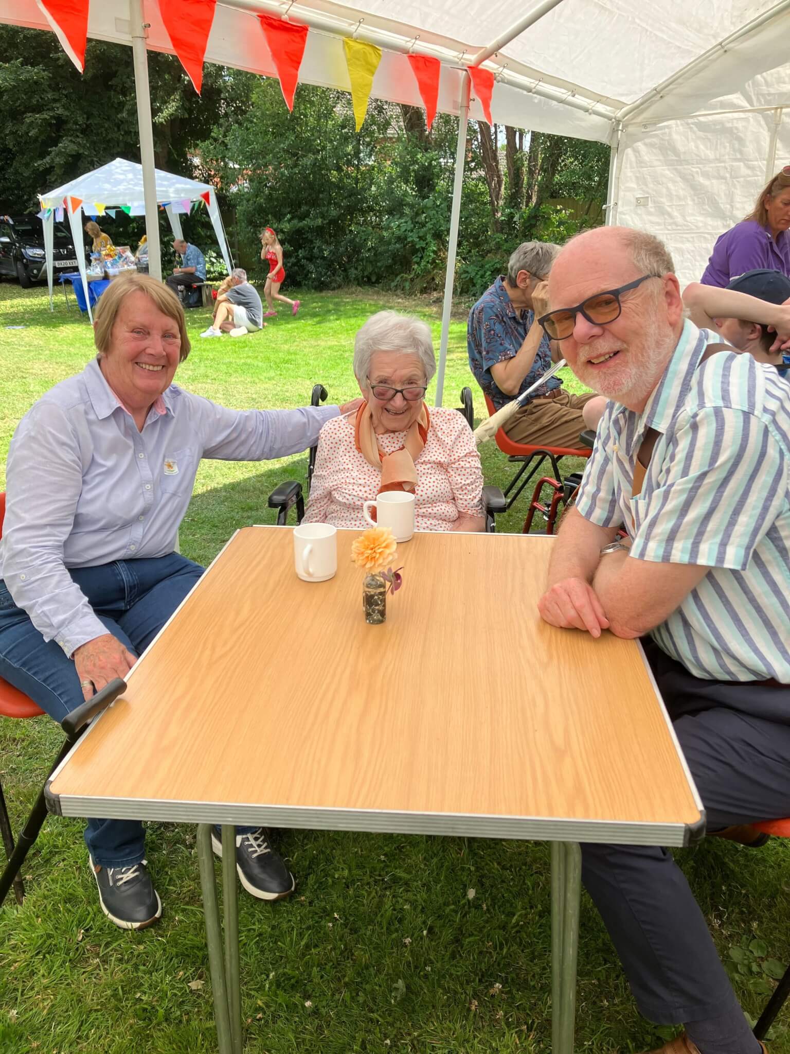 Three elderly individuals sitting at a wooden table under a canopy, enjoying a social event outdoors. - Home Instead