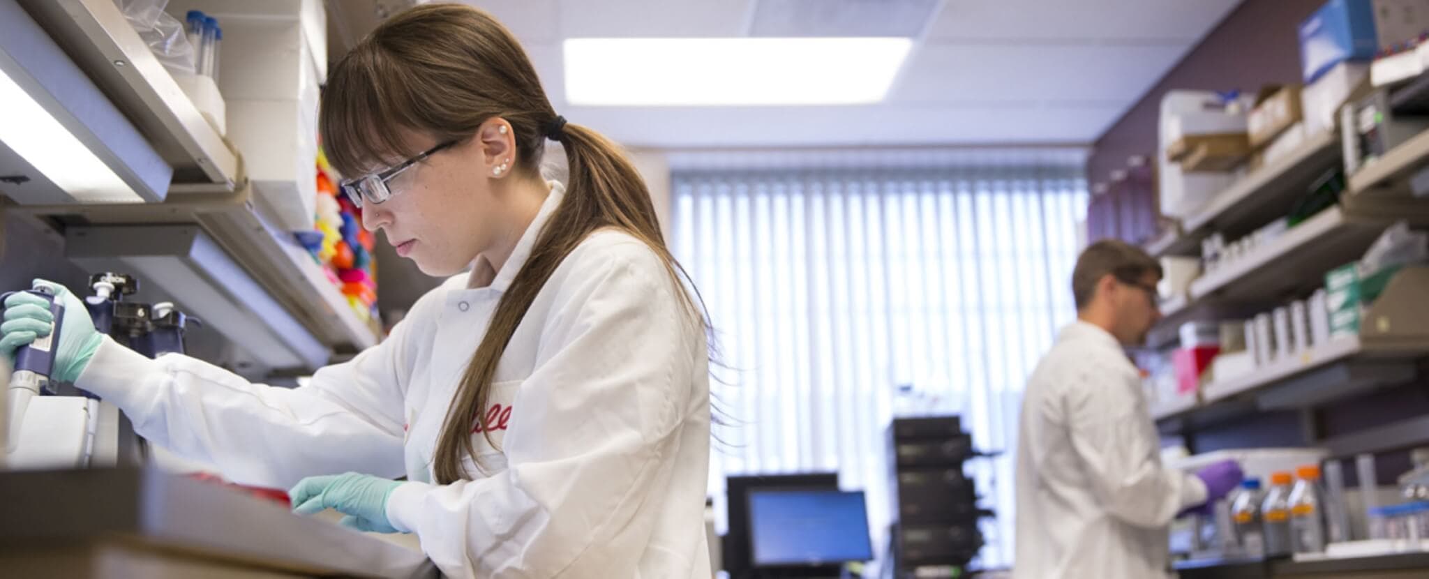 Two scientists in lab coats and safety glasses working in a laboratory with various equipment and supplies. - Home Instead