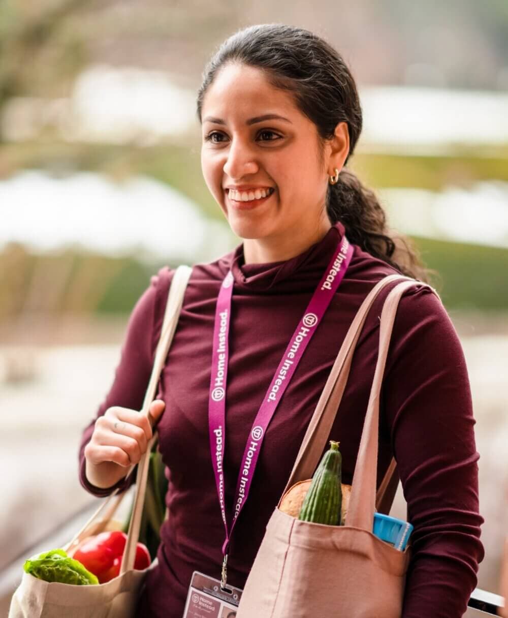 Person smiling while carrying grocery bags, wearing a lanyard with "home instead" text, outdoors. - Home Instead