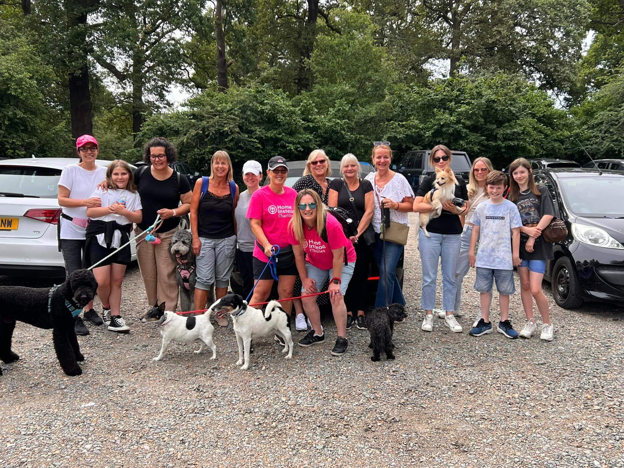 Group of 13 people and several dogs posing outdoors in a parking lot, surrounded by cars and trees. - Home Instead