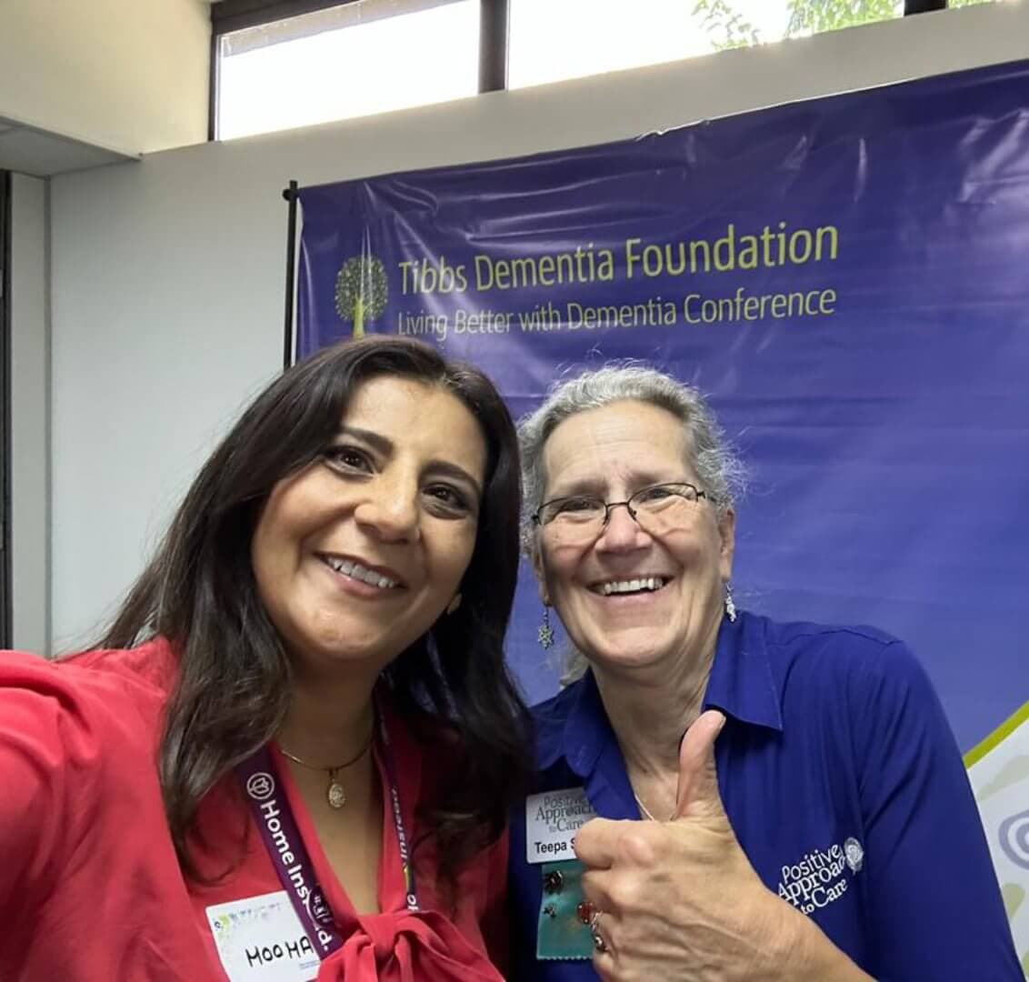 Two women smiling in front of a Tibbs Dementia Foundation banner at a conference. One woman is giving a thumbs-up. - Home Instead