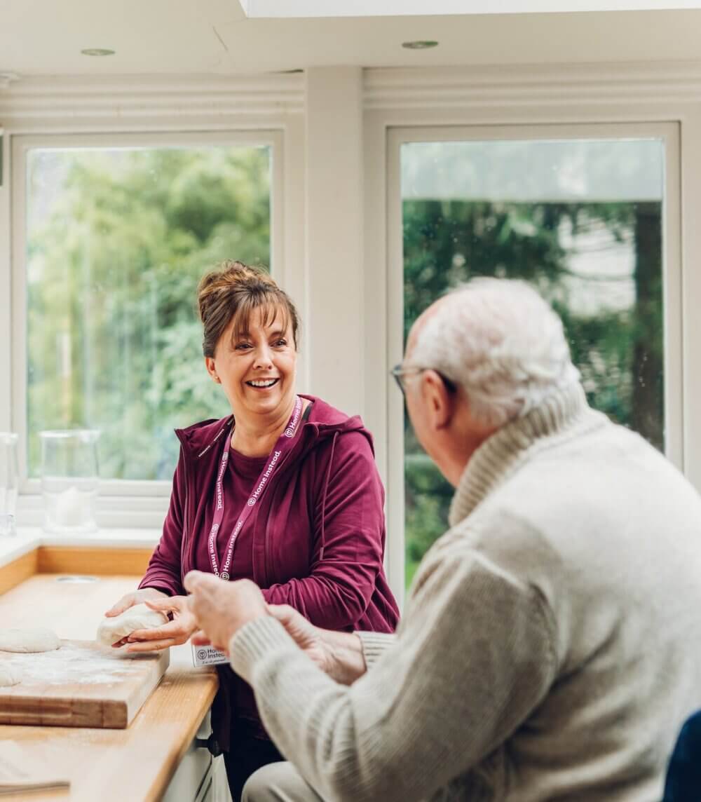 A woman and an elderly man sit at a table, smiling, as they look at a tablet device together. - Home Instead