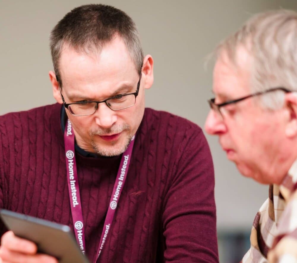 Two men, one wearing a 'Home Instead' lanyard, look at a tablet together in a close-up shot. - Home Instead
