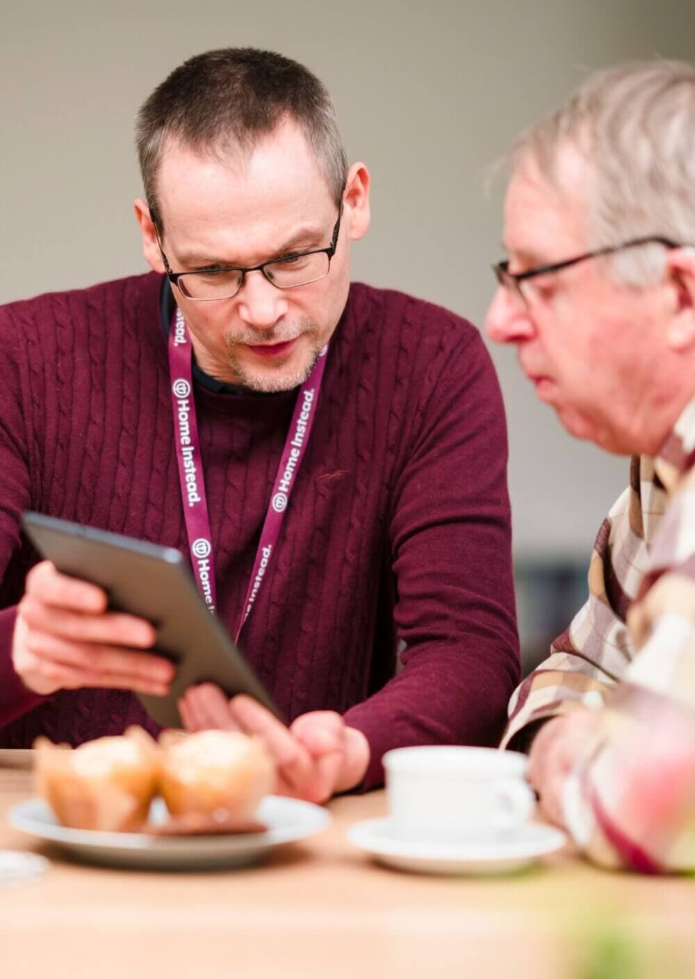 Two men at a table, one showing the other a tablet. There's coffee and pastries on the table. - Home Instead