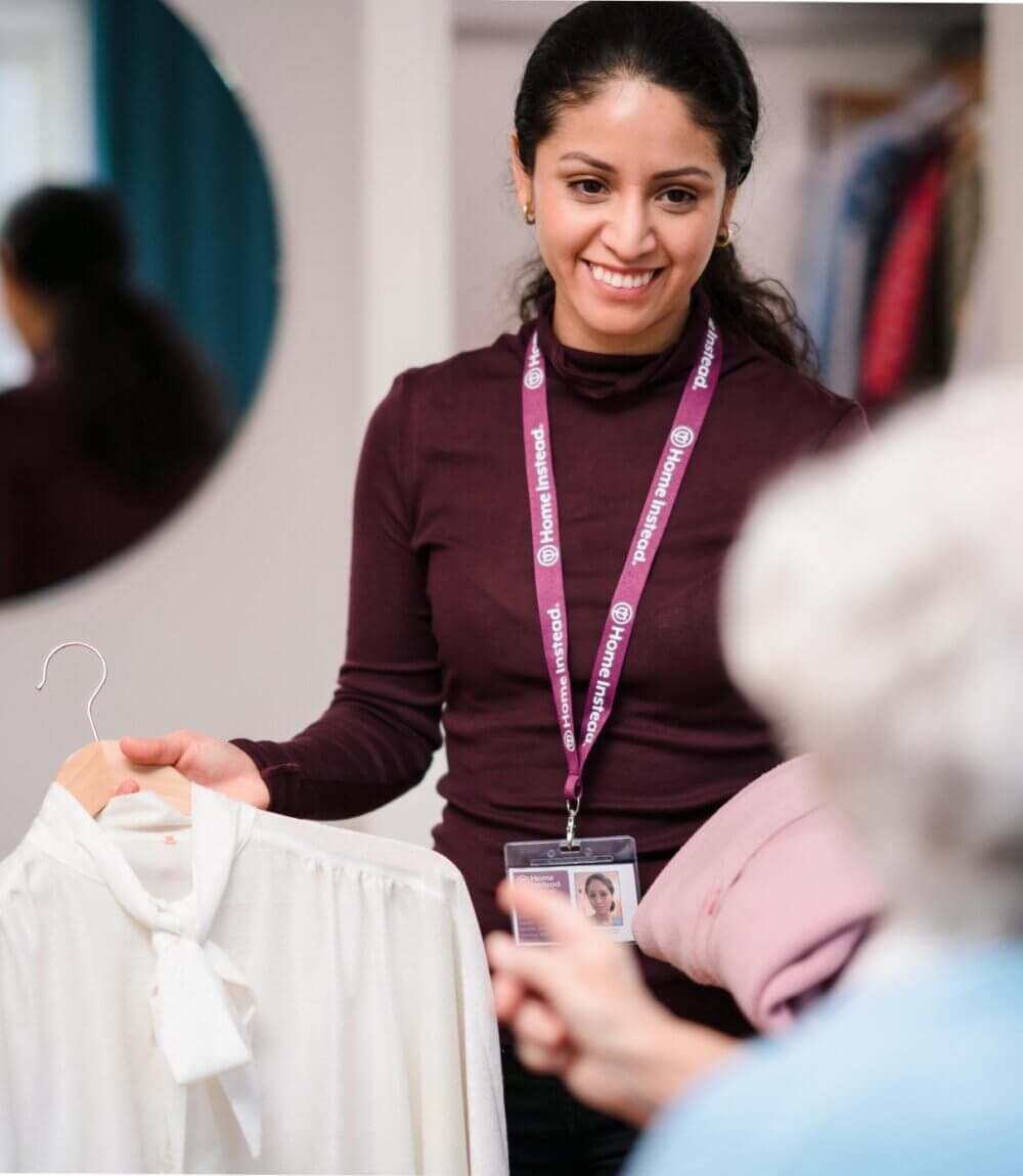 A woman assists an older person with clothing choices in a room with a mirror, holding a white blouse on a hanger. - Home Instead