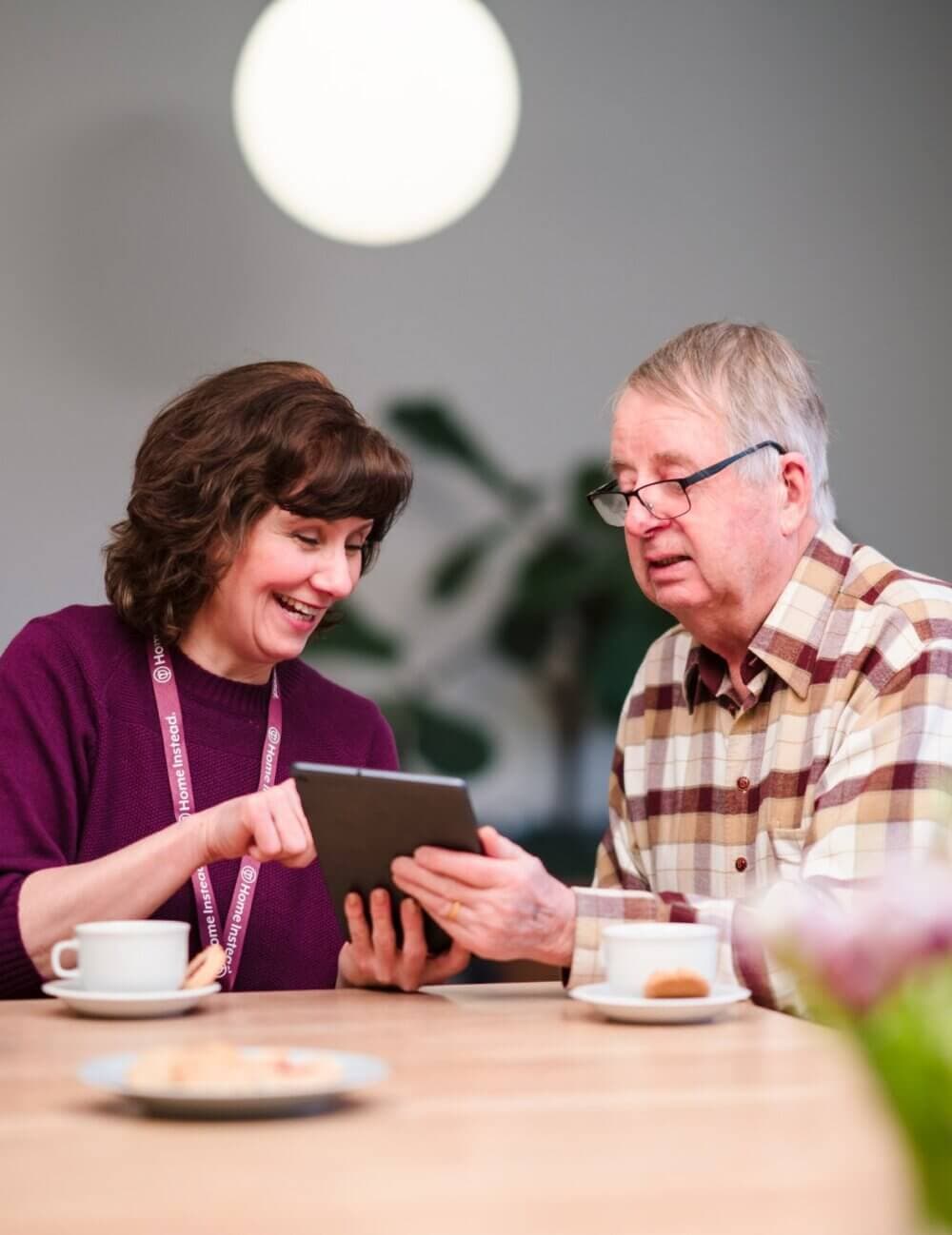 A woman showing a man something on a tablet at a table with coffee cups and plates. - Home Instead