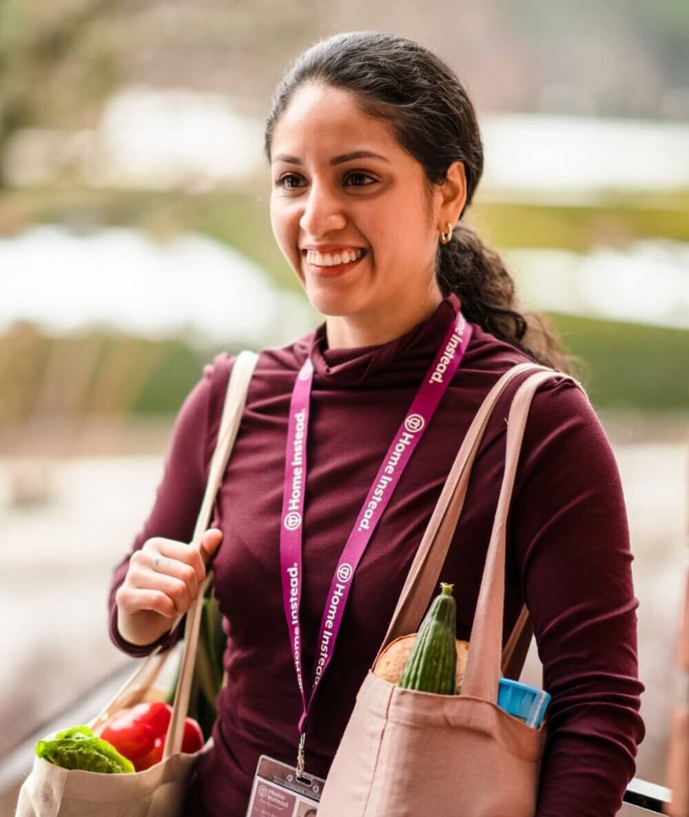 Woman holding dog and elderly lady smiling at another woman carrying groceries at the door. - Home Instead