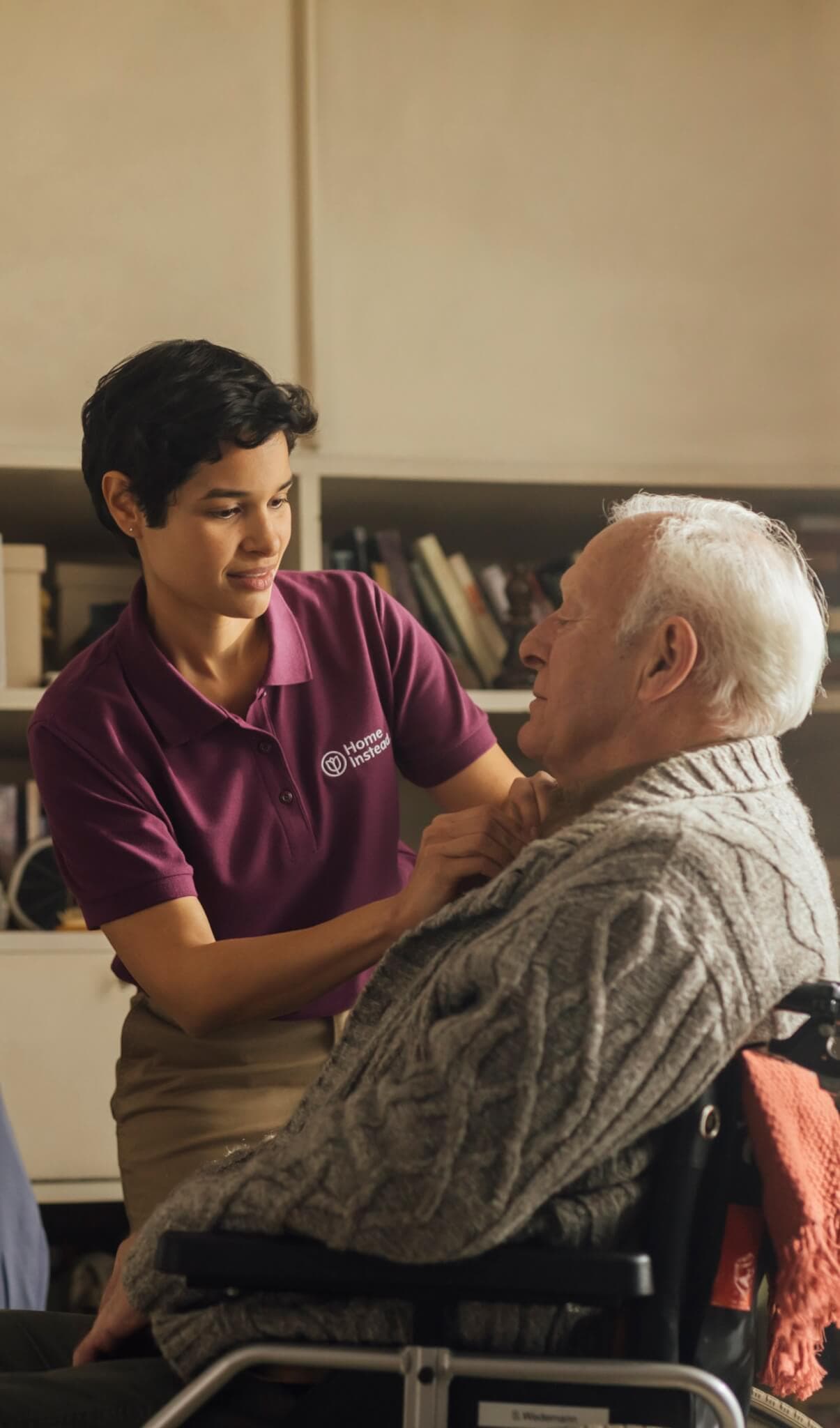 A caregiver in a maroon shirt assists an elderly person in a wheelchair inside a warmly lit room. - Home Instead