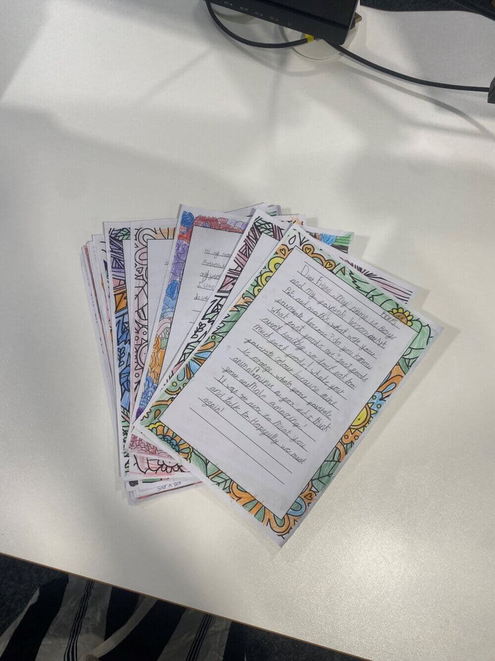 A stack of handwritten letters with colorful, intricately decorated borders on a white table. - Home Instead