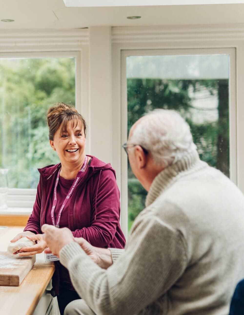 Caregiver in maroon top and lanyard smiles while talking to an elderly man in glasses and a grey sweater at a table. - Home Instead