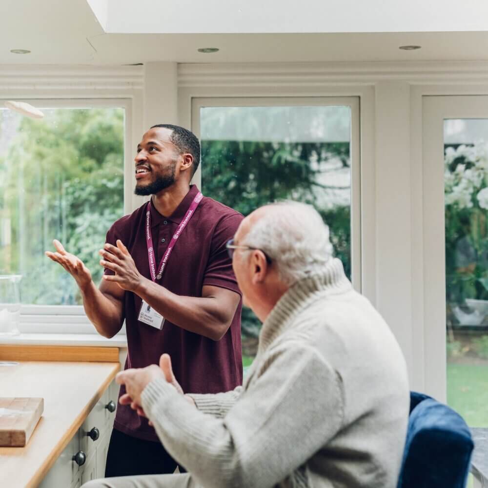 A caregiver in a maroon shirt and name tag speaks with an elderly man in a white sweater in a bright room. - Home Instead