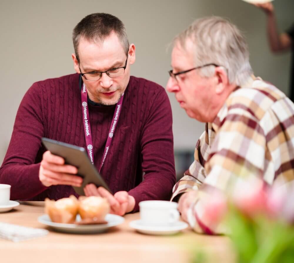 Two men sitting at a table, one in a maroon sweater showing a tablet to the other in a plaid shirt, muffins and coffee nearby. - Home Instead