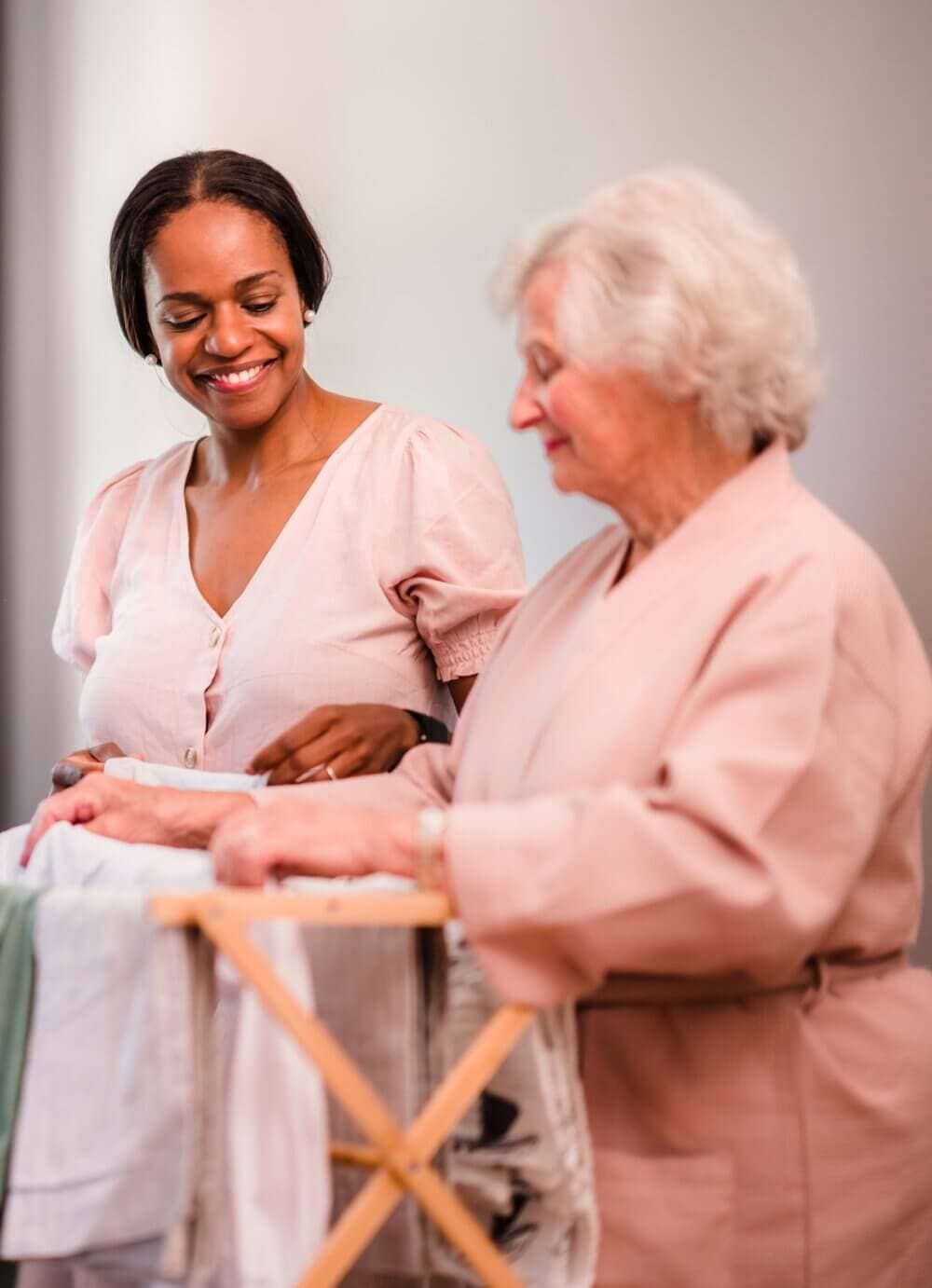 A young woman and an elderly woman smile while working together on a laundry task indoors. - Home Instead