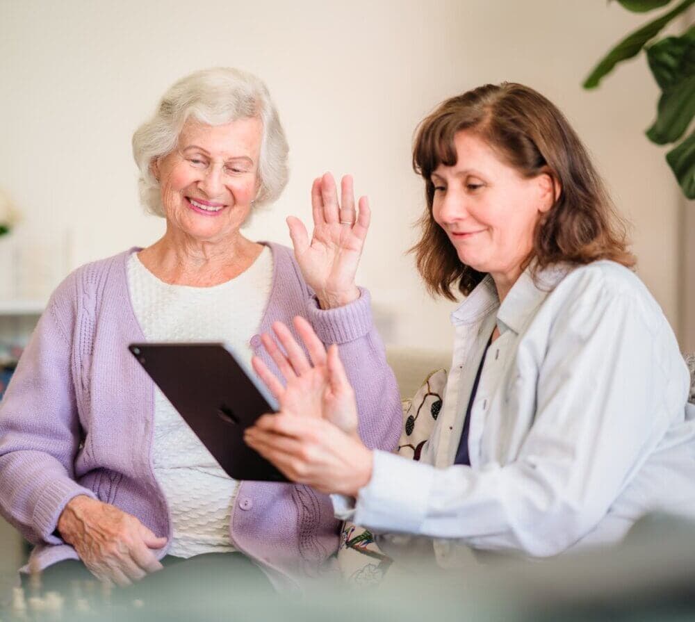 Younger woman smiling at an elderly woman as they fold laundry together in a bright room. - Home Instead