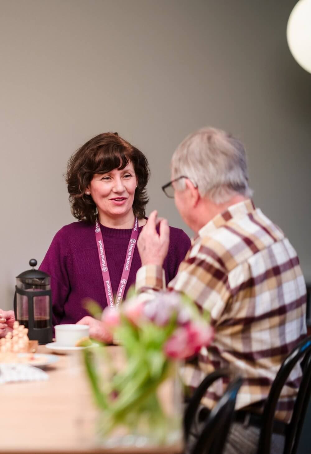 Two people sitting at a table, engaging in conversation; one wears a lanyard, and there is a French press and flowers on the table. - Home Instead