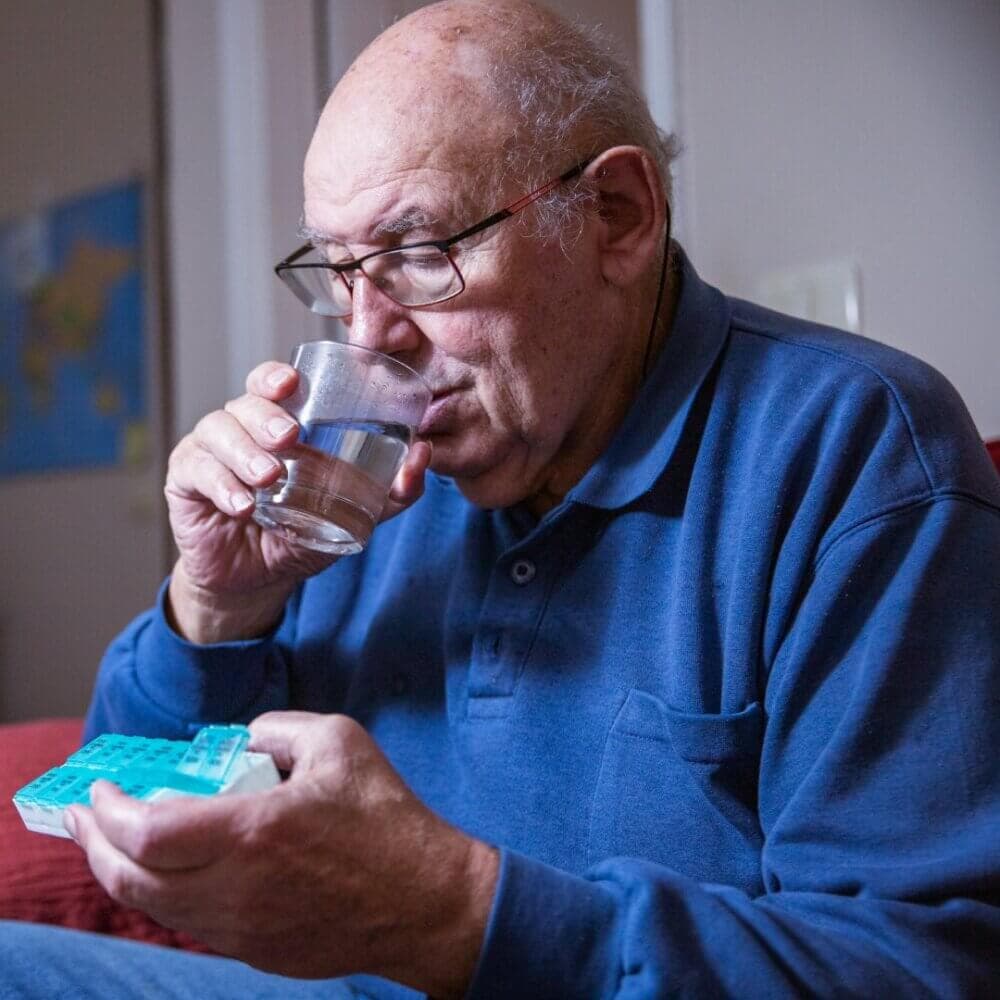 An elderly man drinks a glass of water while holding a pill organizer, seated in a room. - Home Instead