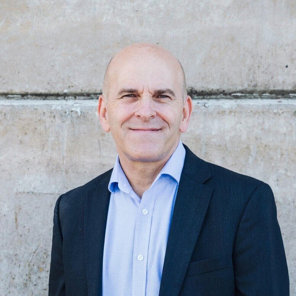 Smiling bald man in a suit and light blue shirt stands in front of a concrete wall. - Home Instead