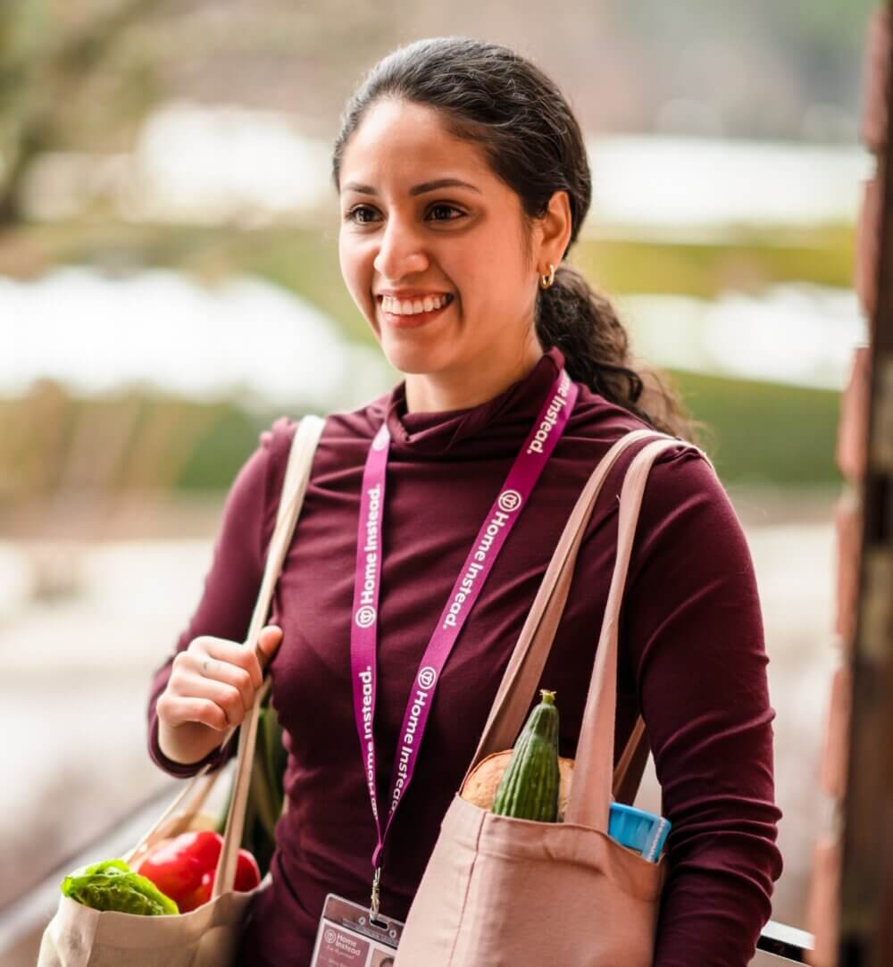 A smiling woman wearing a maroon top and lanyard, holding grocery bags with vegetables, standing outdoors. - Home Instead