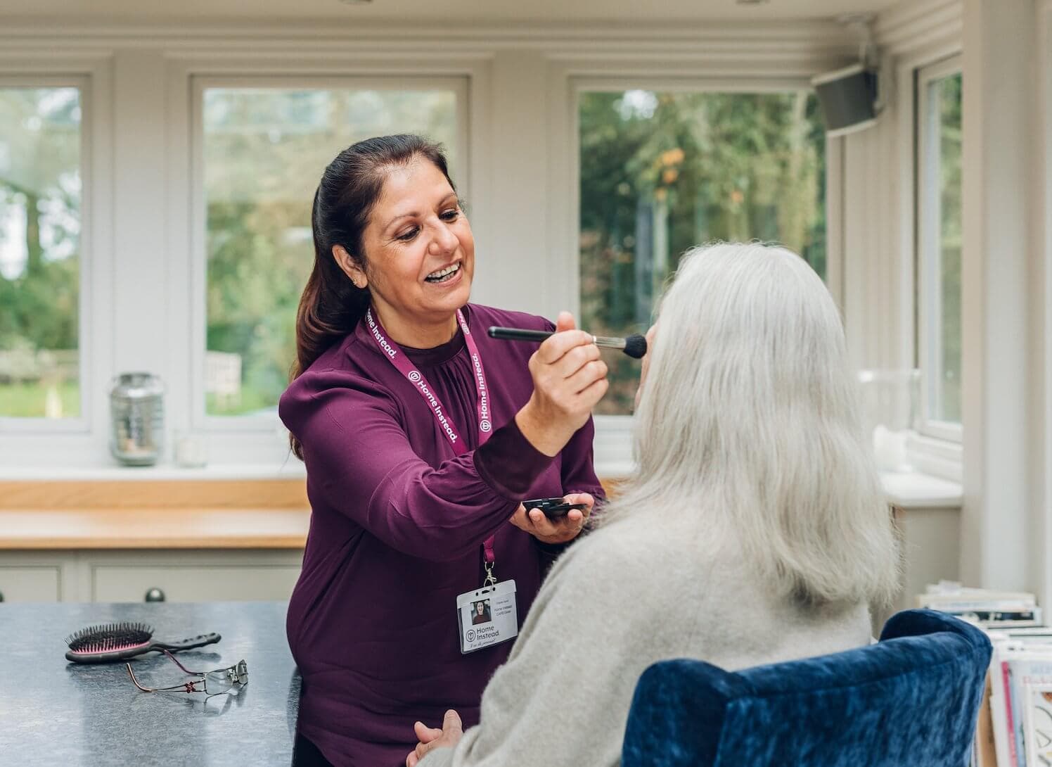 A caregiver smiles while applying makeup to an elderly woman seated in a cozy, well-lit room. - Home Instead